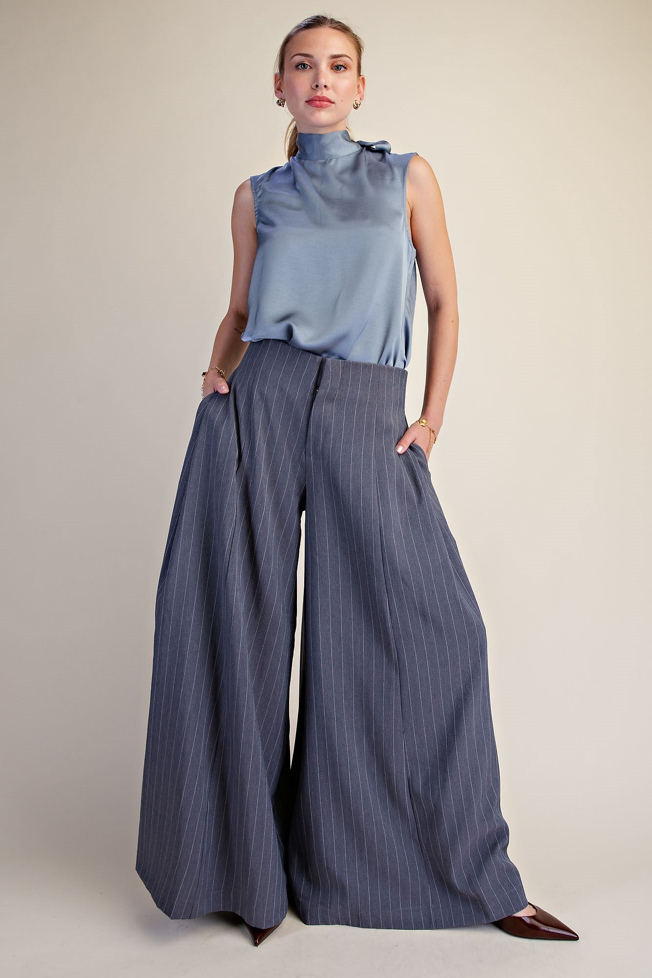 A model wearing a sleeveless light blue mock-neck top and wide-leg pinstripe trousers in a slate blue-gray color, paired with brown pointed-toe flats, against a plain white background.