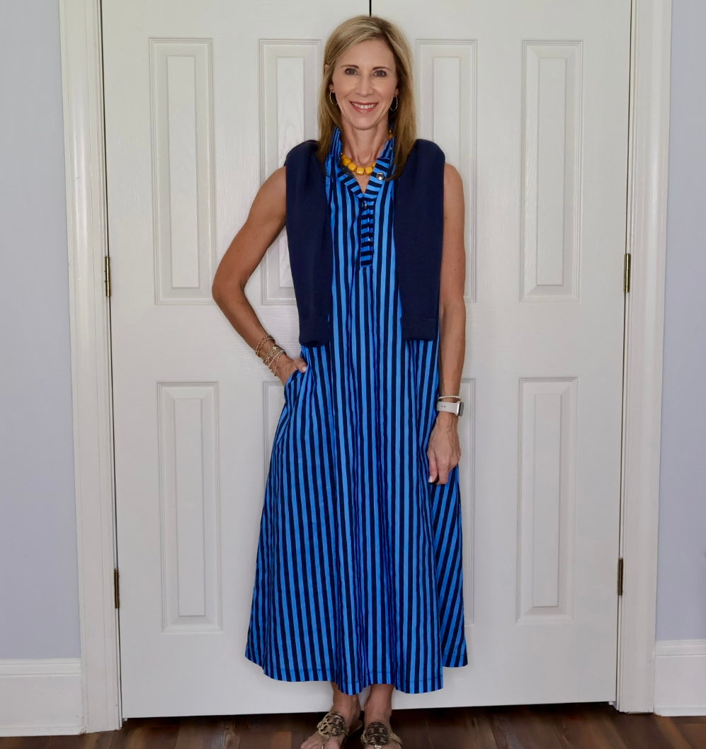 A woman stands in front of white paneled doors wearing a sleeveless blue and white vertically striped midi dress with a navy sweater, a chunky gold necklace, and gold print shoes.
