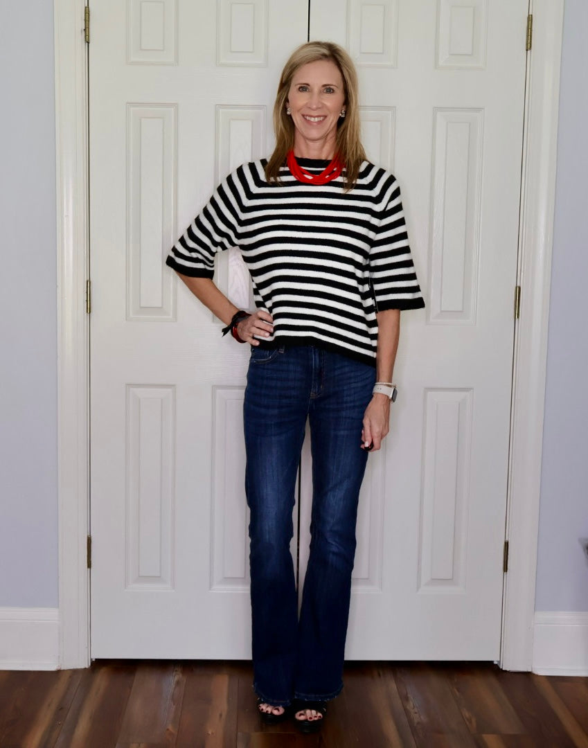 A woman stands in front of white paneled doors wearing a black and white horizontally striped short-sleeve top, dark blue jeans, and a bright red necklace.