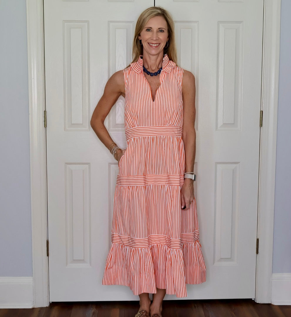A woman stands in front of a white paneled door wearing a sleeveless tiered maxi dress with vertical orange and white stripes, a split neckline, and a ruffled hem.