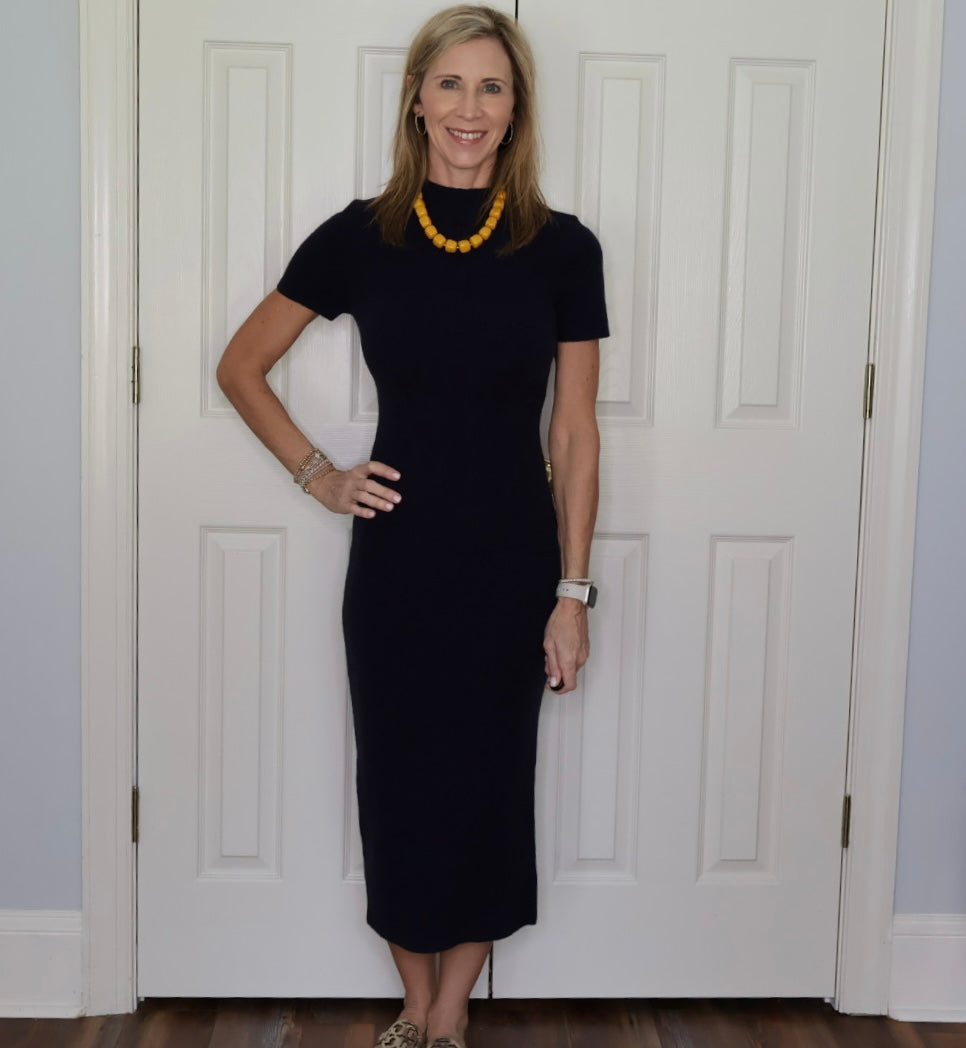 A woman wearing a dark navy
short-sleeve midi dress and a chunky yellow beaded necklace stands in front of white paneled doors.
