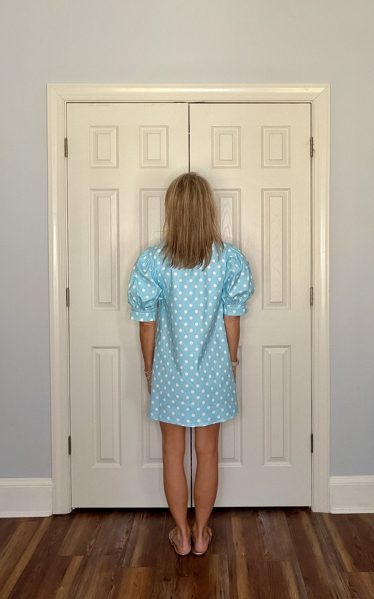Person wearing a blue polka dot dress standing in front of a white door.