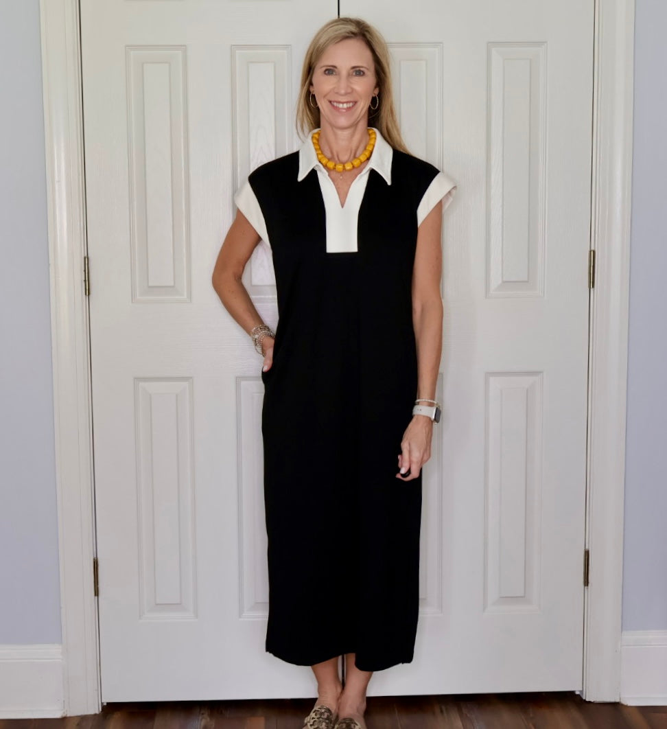 A woman modeling a black midi dress with a white collar and short white sleeves, standing in front of white paneled doors.