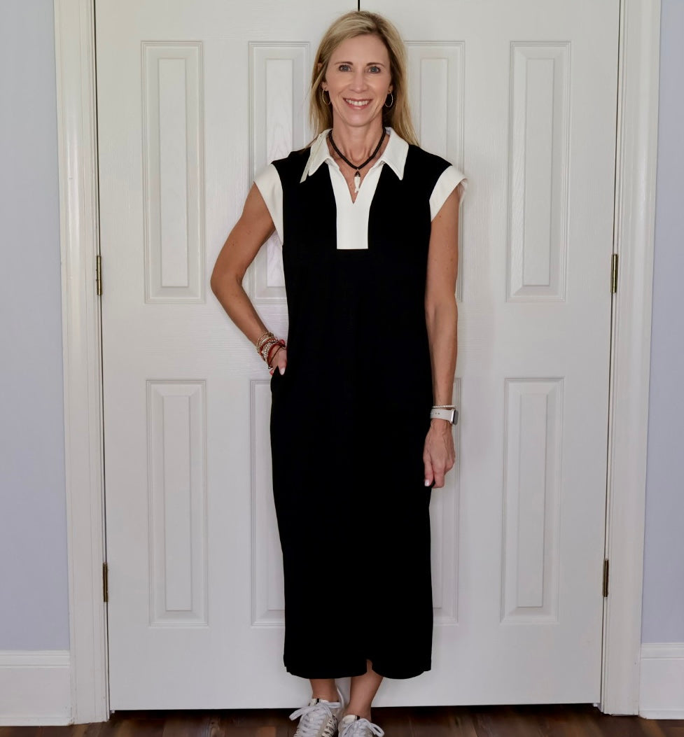 A woman stands in front of a white paneled door wearing a black sleeveless maxi dress with a white collar and placket, paired with white sneakers.