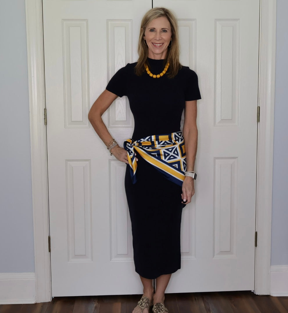 A woman stands in front of white paneled doors wearing a dark navy short-sleeve midi dress with a yellow, blue, and white patterned scarf tied around her waist. She is also wearing a yellow beaded necklace and leopard print heels.