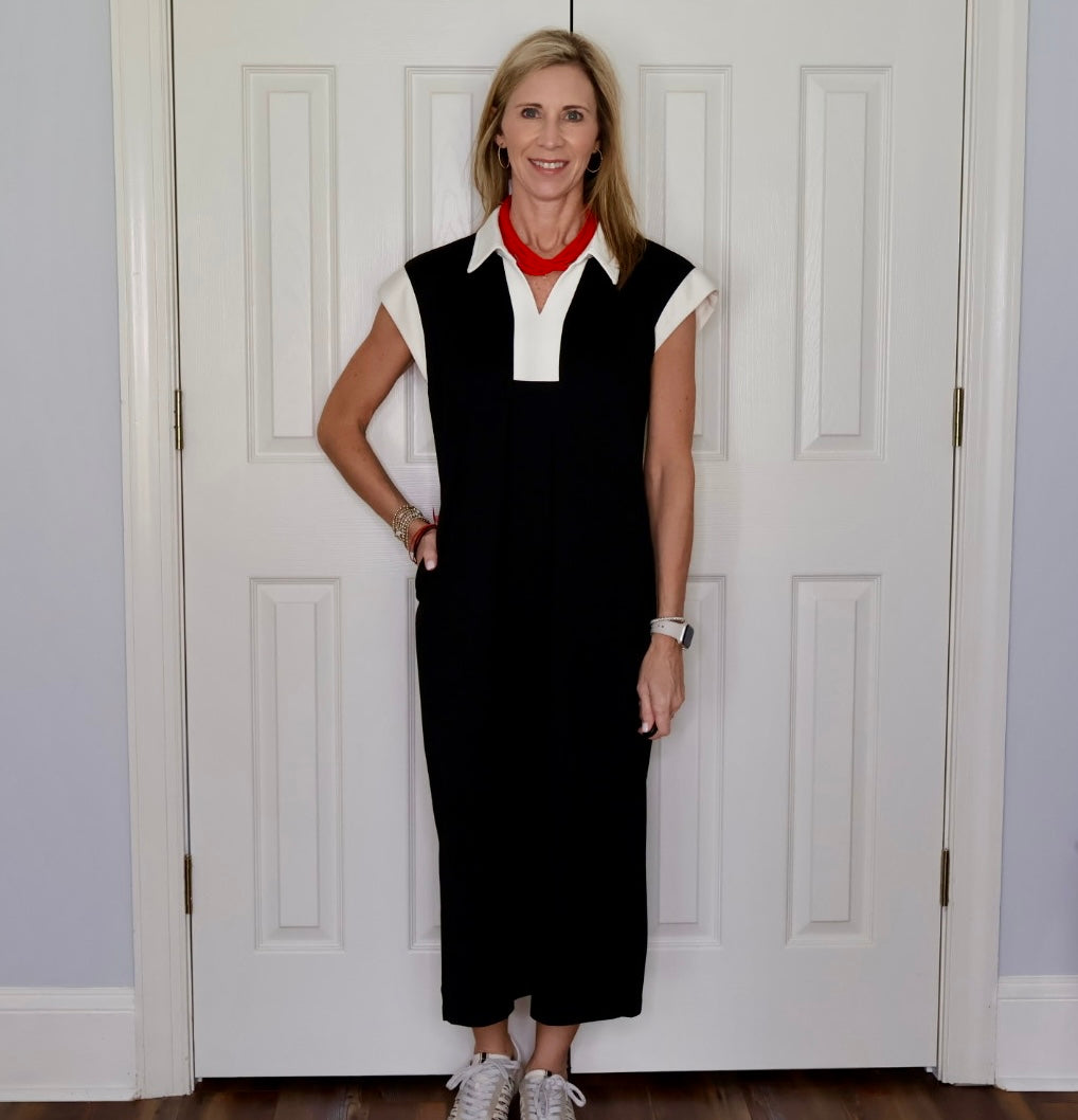 A woman stands in front of white paneled doors wearing a black sleeveless midi dress with a white collar and white shoulder panels, a bright red neck scarf, and white sneakers.