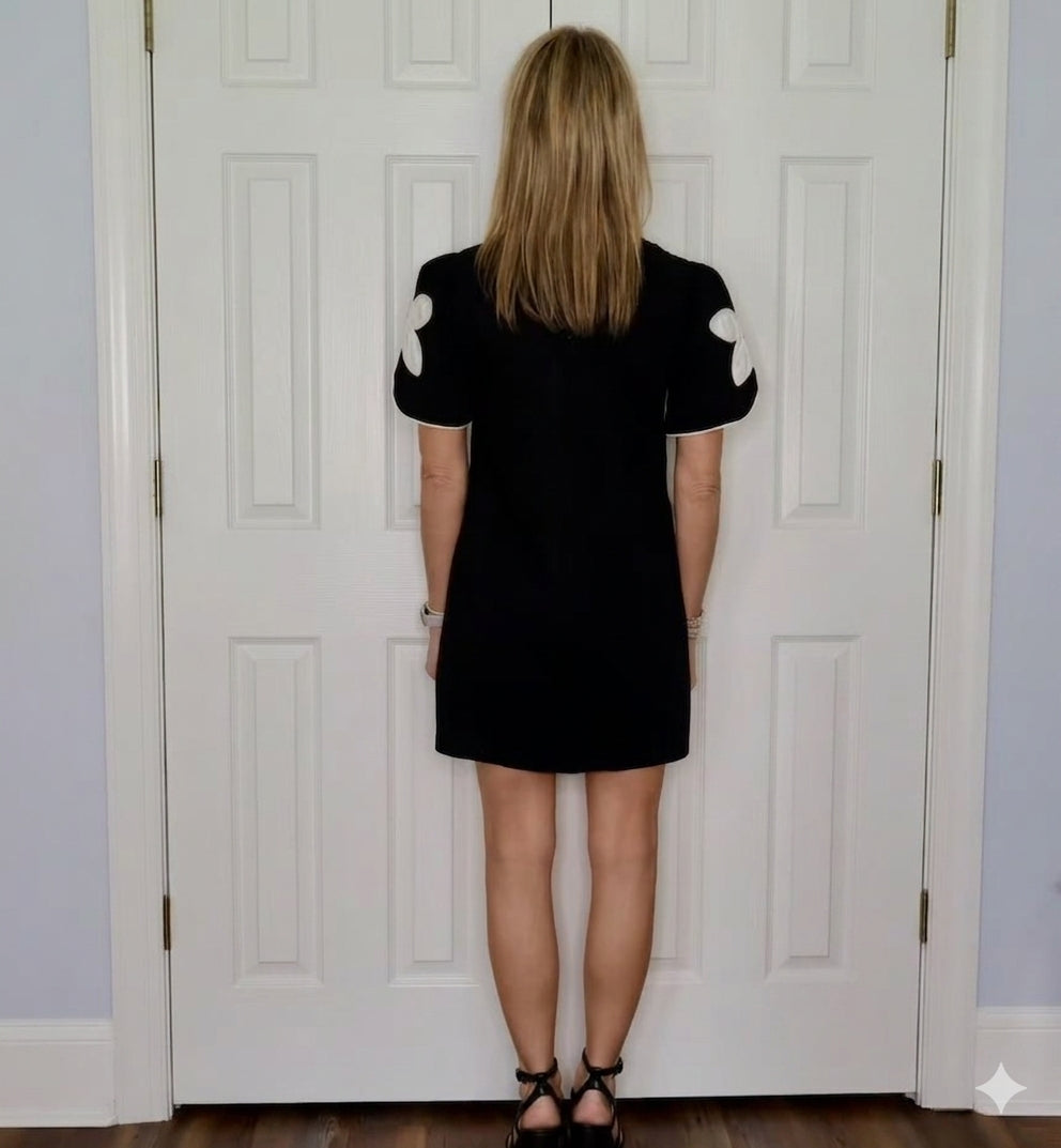 A woman stands with her back to the camera in front of white paneled doors, wearing a short black dress with white floral applique on the sleeves and black strappy heels.