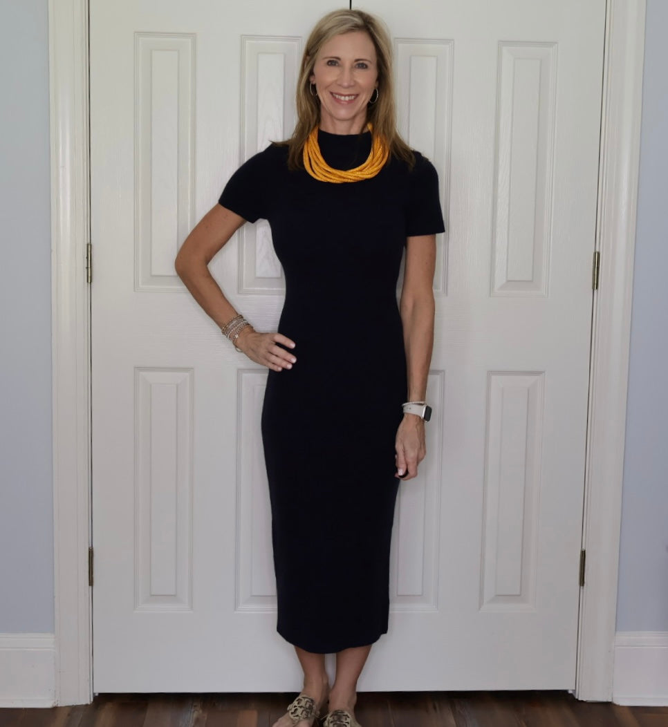 A woman stands in front of a white paneled door wearing a dark navy short-sleeve midi dress, a chunky yellow necklace, bracelets, and gold print shoes.