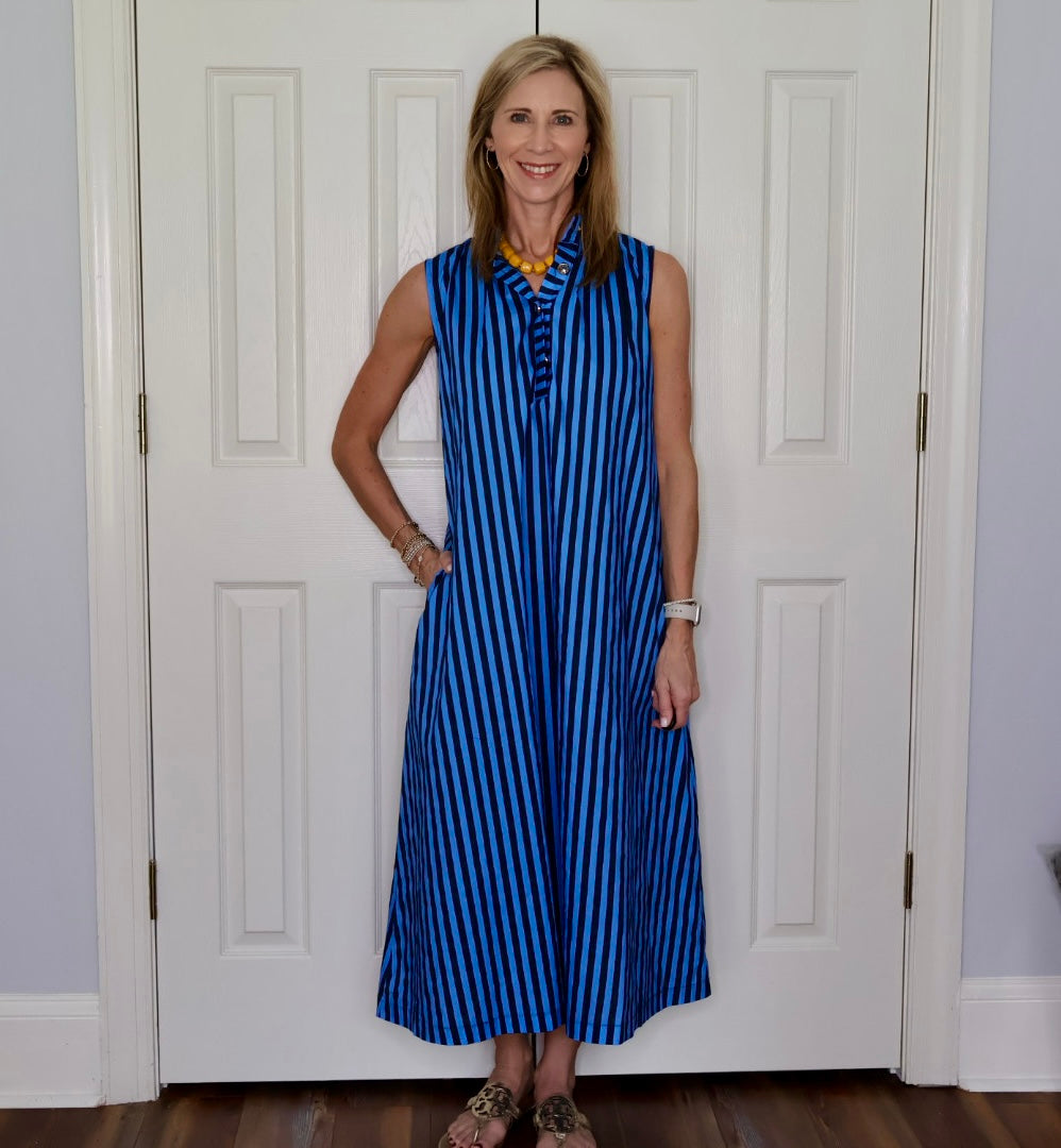 A woman stands in front of white paneled doors wearing a sleeveless blue and black vertically striped maxi dress with a buttoned neckline and pockets.
