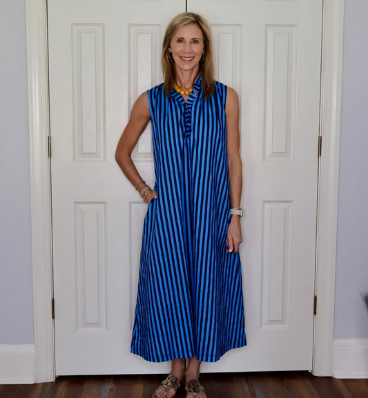A woman stands in front of white paneled doors wearing a sleeveless blue and black vertically striped maxi dress with a buttoned neckline and pockets.