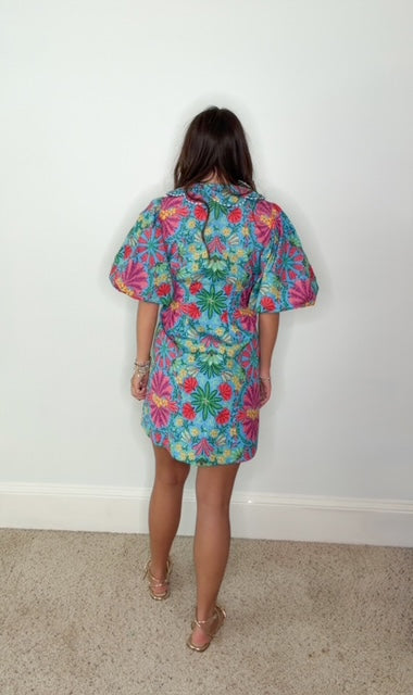 A woman viewed from behind wearing a short-sleeve mini dress with a colorful floral print on a blue background, standing on a carpeted floor against a plain white wall.