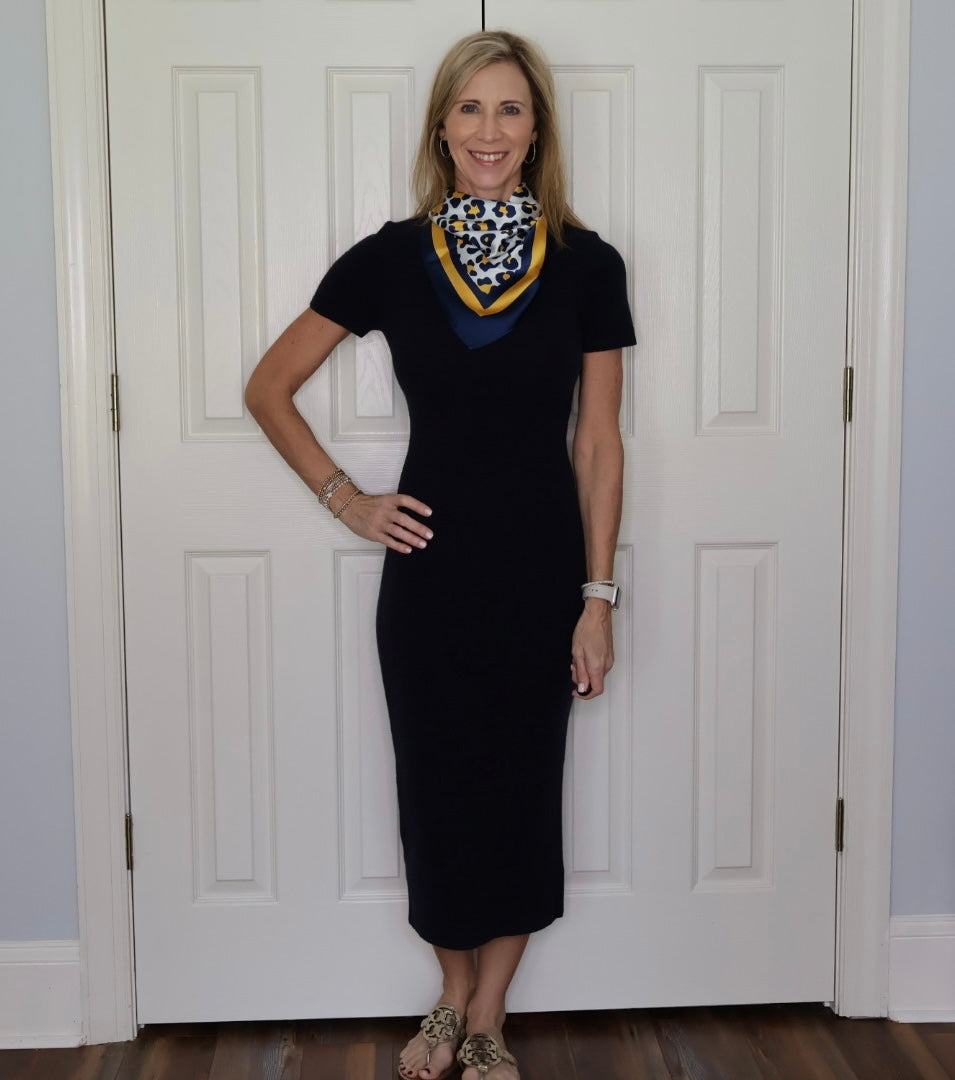 A woman stands in front of white paneled doors wearing a dark navy short-sleeve midi dress styled with a blue, yellow, and black patterned neck scarf.