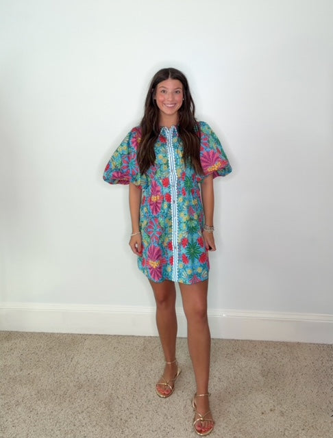 A young woman stands against a plain white wall wearing a short, colorful floral print dress with puff sleeves and a white vertical trim down the front, paired with tan strappy sandals.