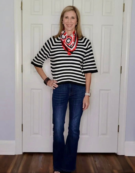 A woman modeling a black and white horizontally striped short-sleeve knit top with a red, white, and black paisley bandana tied around her neck, paired with dark blue jeans, standing in front of white interior doors.