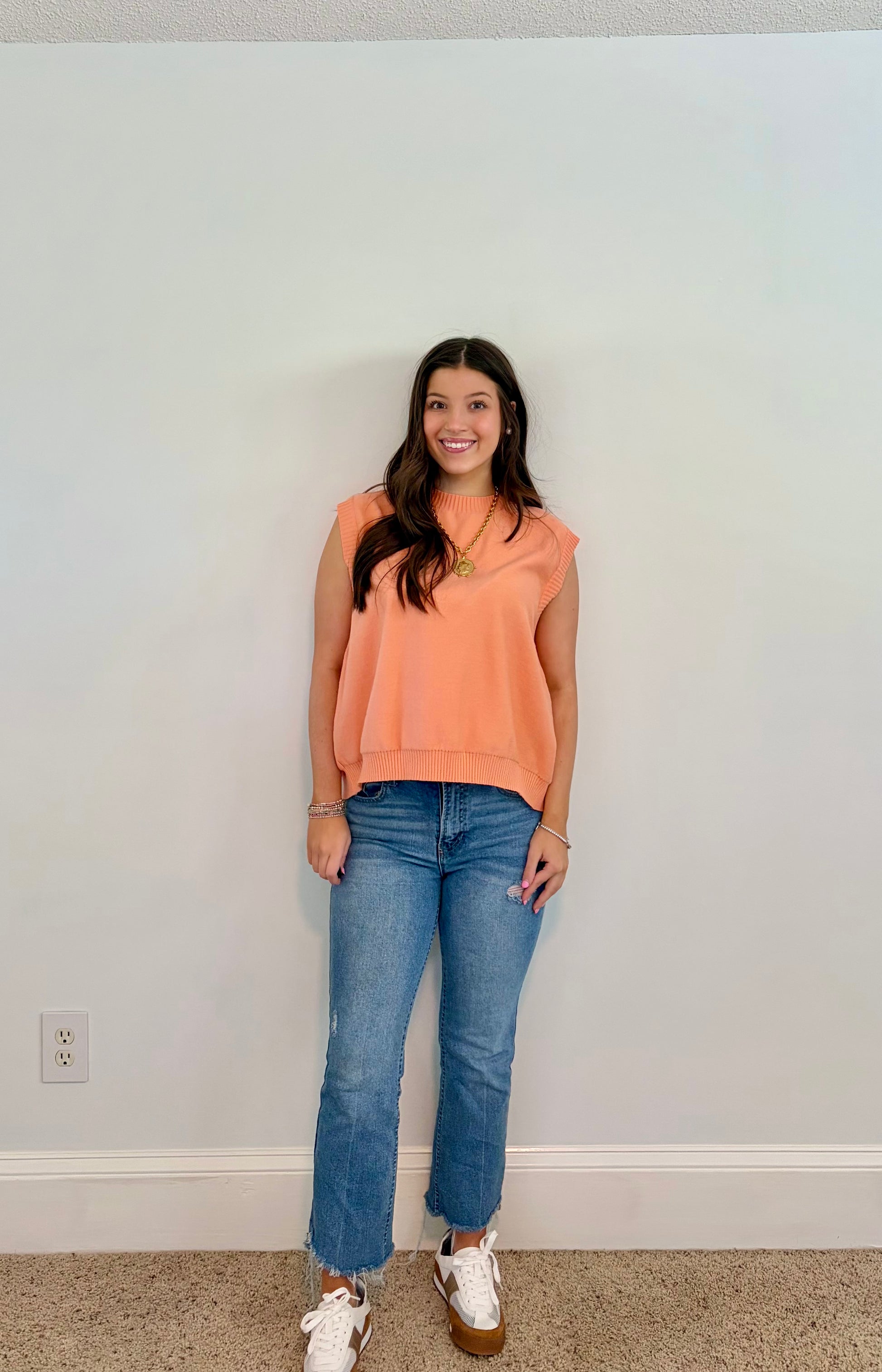 Woman wearing a peach sleeveless top and blue jeans standing against a plain wall.