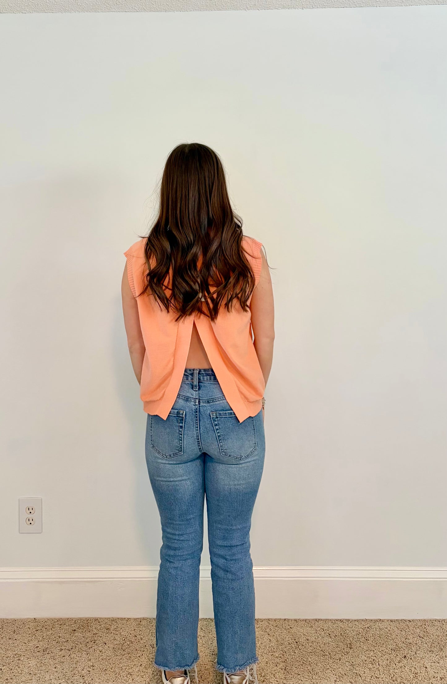 Person wearing a peach tank top and blue jeans standing against a plain wall.