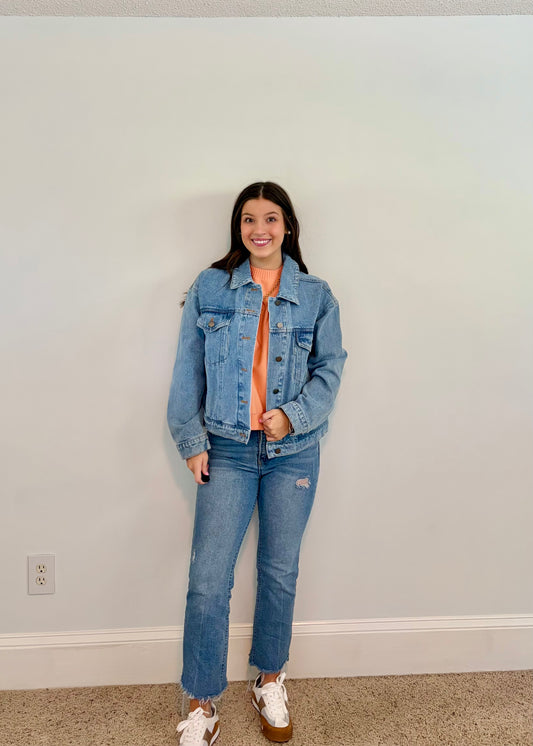 Person wearing a denim jacket and jeans standing against a plain wall.