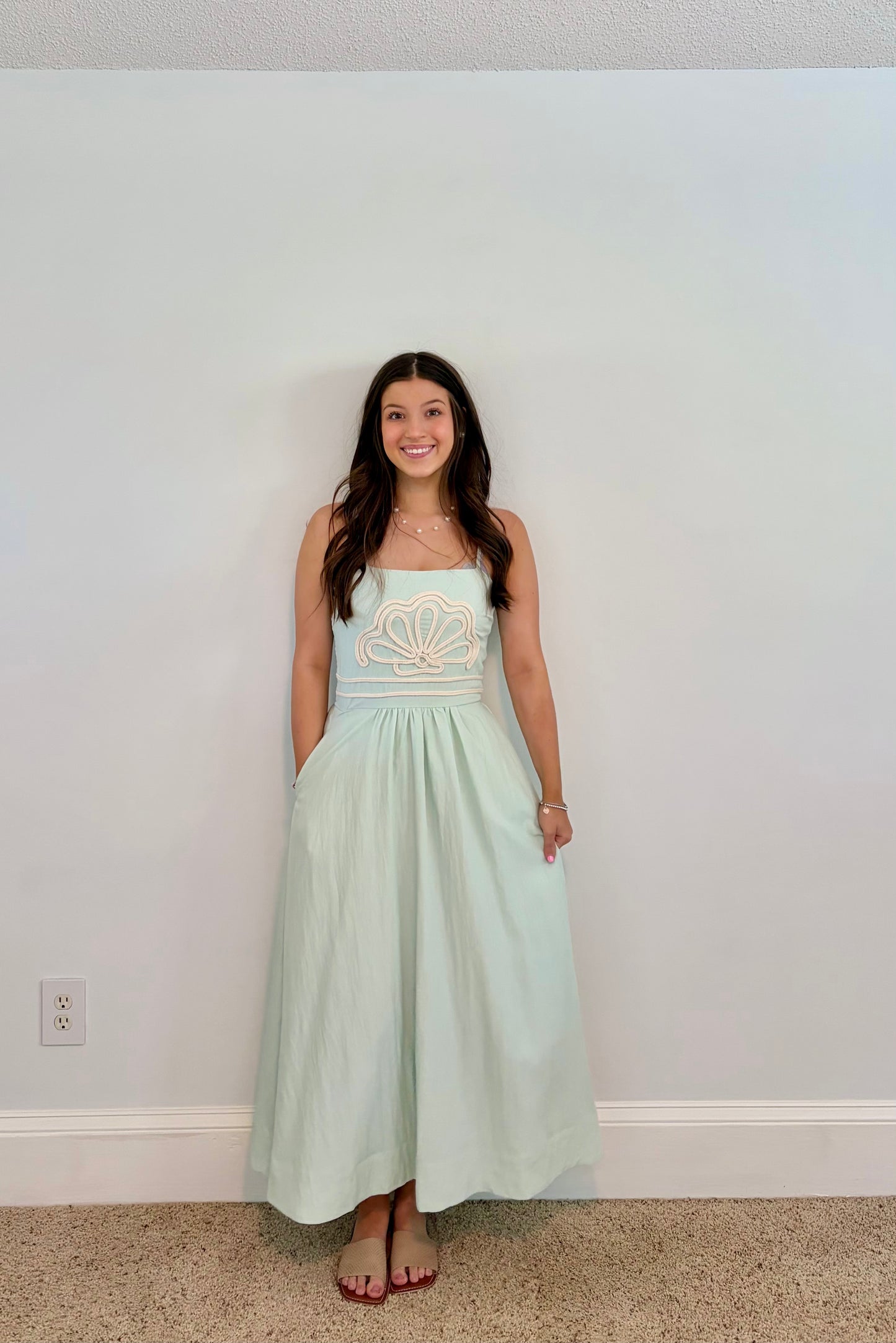 Woman wearing a light green dress with shell  embroidery against a plain wall.
