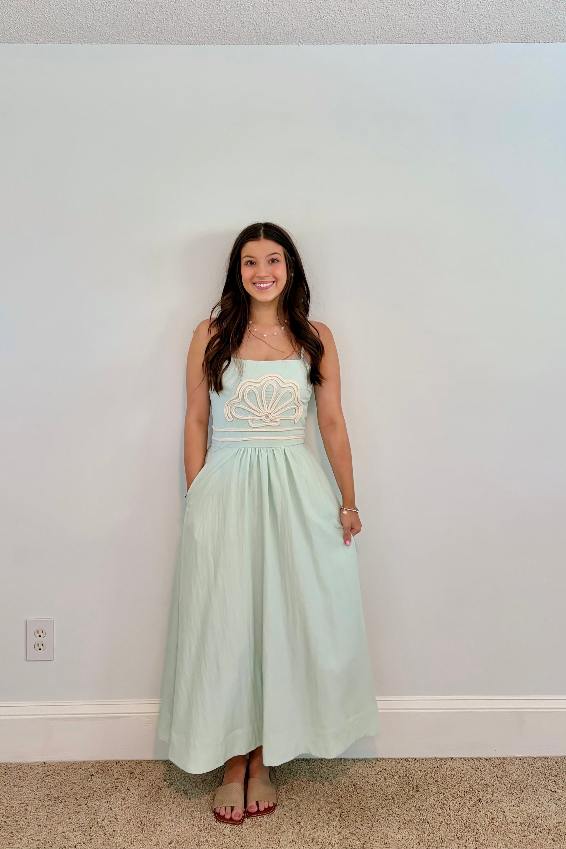 Woman wearing a light green dress with shell  embroidery against a plain wall.