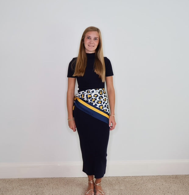 A young woman stands against a plain white wall wearing a dark navy mock-neck short-sleeve fitted midi dress with a diagonal wrap-style scar featuring a white leopard print and navy and yellow stripes.