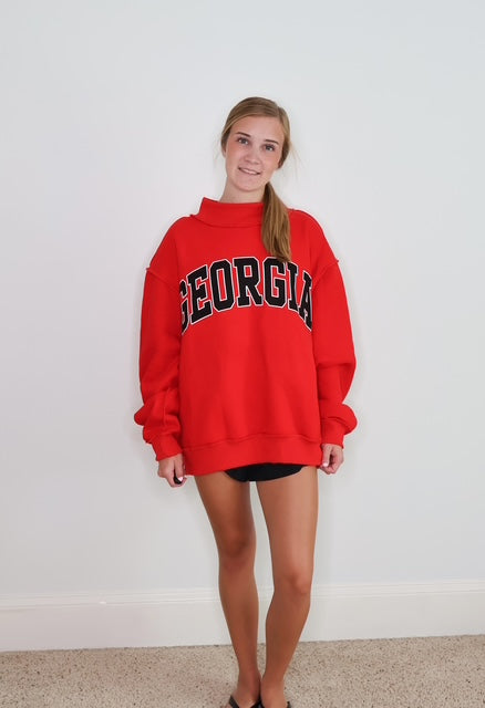 A woman modeling a bright red sweatshirt with 'GEORGIA' printed across the chest in black and white block letters, standing against a plain white wall.