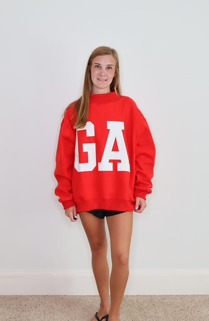 A woman modeling a bright red crewneck sweatshirt with large white block letters 'GA' on the front, paired with black shorts, standing against a plain white wall.