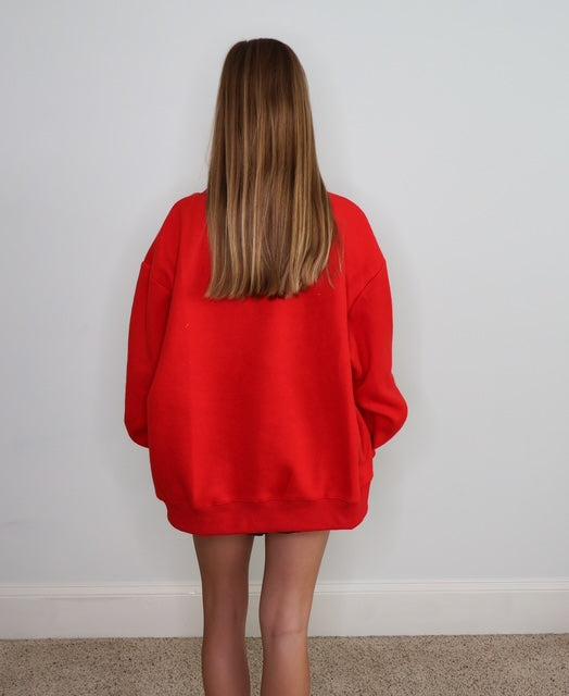 A person viewed from behind wearing an oversized bright red sweatshirt, standing against a plain light gray wall on a beige carpeted floor.
