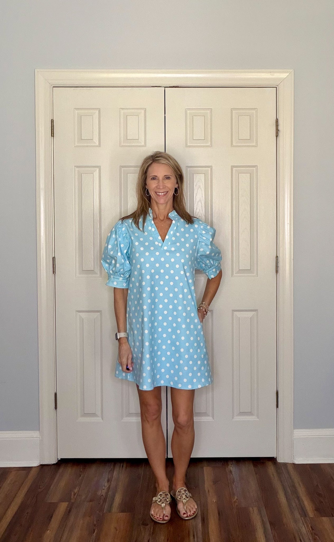 Woman wearing a light blue polka dot dress standing in front of a white door.