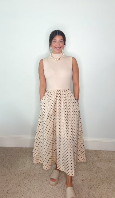 A woman modeling a sleeveless beige mock-neck top and a matching beige midi skirt with small black polka dots, paired with a chunky pearl necklace and beige slide sandals, standing against a plain white wall.