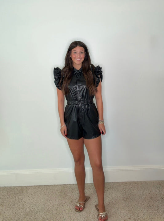 A young woman stands against a plain white wall wearing a shiny black faux-leather romper with ruffled cap sleeves paired with gold strap sandals.
