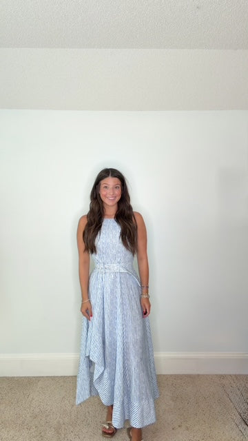 A woman wearing a sleeveless light blue and white vertically striped midi dress with a matching tie belt, standing against a plain white wall.