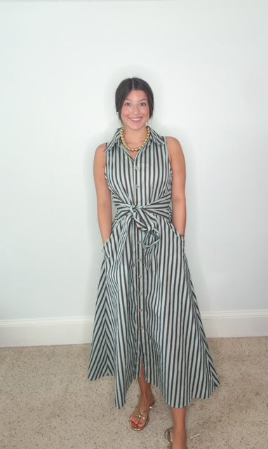 A woman modeling a sleeveless midi dress with vertical seafoam green and chocolate brown stripes, featuring a tie waist, button front, and side pockets, standing against a plain white wall.